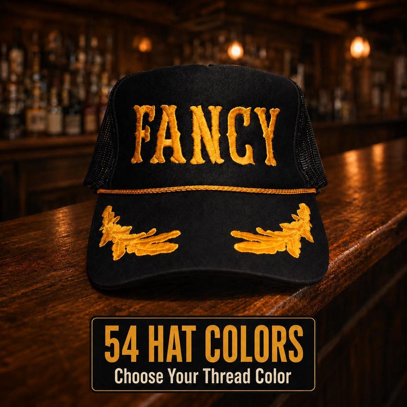May include: A black trucker hat featuring the word FANCY embroidered in gold thread across the front panel, with gold laurel leaf embroidery on the brim and a gold cord trim. A rectangular graphic below displays the text 54 HAT COLORS CHOOSE YOUR THREAD COLOR.