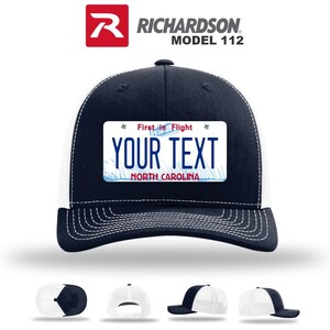 May include: Navy blue and white trucker hat with a license plate design. The hat features a navy blue crown and bill, a white mesh back, and a rectangular patch with the text "YOUR TEXT" and "NORTH CAROLINA". The hat also has the text "First in Flight".