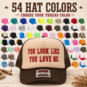 May include: A foam front trucker hat with a brown mesh back and a dark brown brim. The front panel is beige with the red embroidered text "YOU LOOK LIKE YOU LOVE ME". An Otto brand sticker is on the brim. The background displays 54 hat color options.