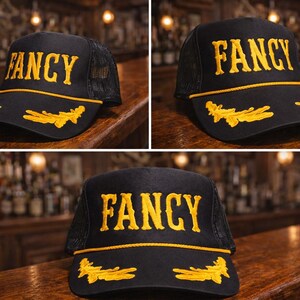 May include: A black trucker hat featuring the word FANCY embroidered in yellow on the front panel. The brim is decorated with yellow embroidered leaf motifs and a yellow cord trim. The back portion of the hat is constructed from black mesh fabric.