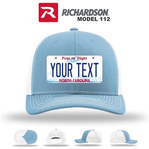 May include: Light blue and white trucker hat with a license plate design. The hat features the text "YOUR TEXT" and "NORTH CAROLINA" on the front. The top of the hat is light blue, with a white mesh back. The Richardson Model 112 logo is visible.