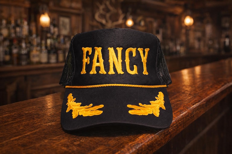 May include: A black trucker hat featuring the word FANCY embroidered in bold yellow letters on the front panel. The cap includes a yellow cord trim above the brim and two yellow embroidered leaf designs on the brim, with black mesh side panels.