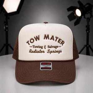 May include: A brown and white trucker hat with the text "TOW MATER - Towing & Salvage - Radiator Springs" embroidered on the front. The hat has a brown mesh back and a brown brim. A small "OTTO" label is on the front.
