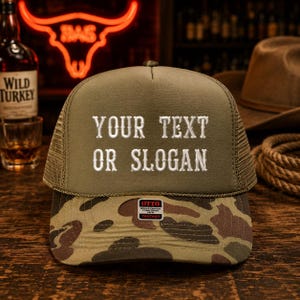 May include: A trucker hat with a green crown and a camouflage pattern on the bill. The hat features the embroidered text "YOUR TEXT OR SLOGAN" in white. The hat has a mesh back and a small tag that says "OTTO WORLD FAMOUS FOAM FRONT TRUCKER".