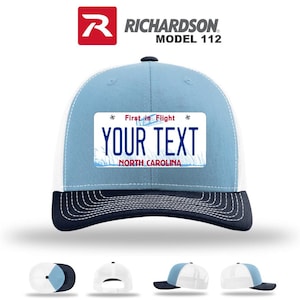 May include: A light blue and white trucker hat with a navy blue brim. The hat features a rectangular patch resembling a North Carolina license plate with the text "YOUR TEXT" and "First in Flight". The brand name "RICHARDSON" is visible.
