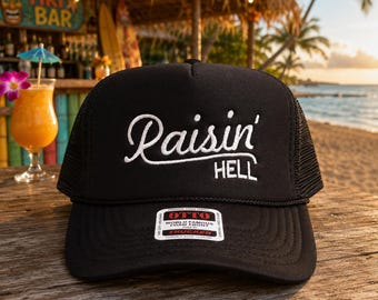 May include: Black trucker hat with the text "Raisin' HELL" in white script. The hat has a mesh back and a curved brim. The hat is sitting on a wooden surface, with a tropical bar and beach in the background.