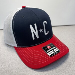 May include: A navy blue and red trucker cap with a white mesh back. The front has the letters "NC" in white, with a star between them. The peak is red, and the cap has a red button on top.