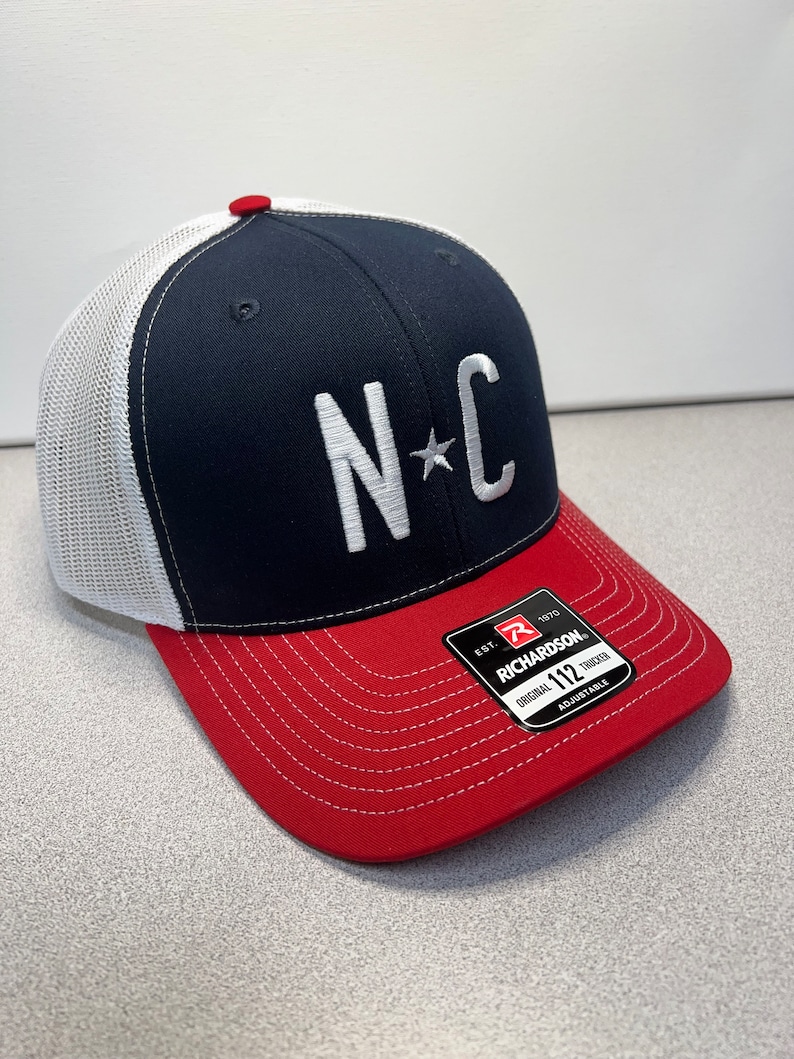 May include: A navy blue and red trucker cap with a white mesh back. The front has the letters "NC" in white, with a star between them. The peak is red, and the cap has a red button on top.