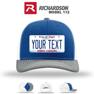 May include: A blue and white trucker hat with a gray brim. The hat features a license plate design with the text "YOUR TEXT" and "NORTH CAROLINA". The hat is a Richardson Model 112.