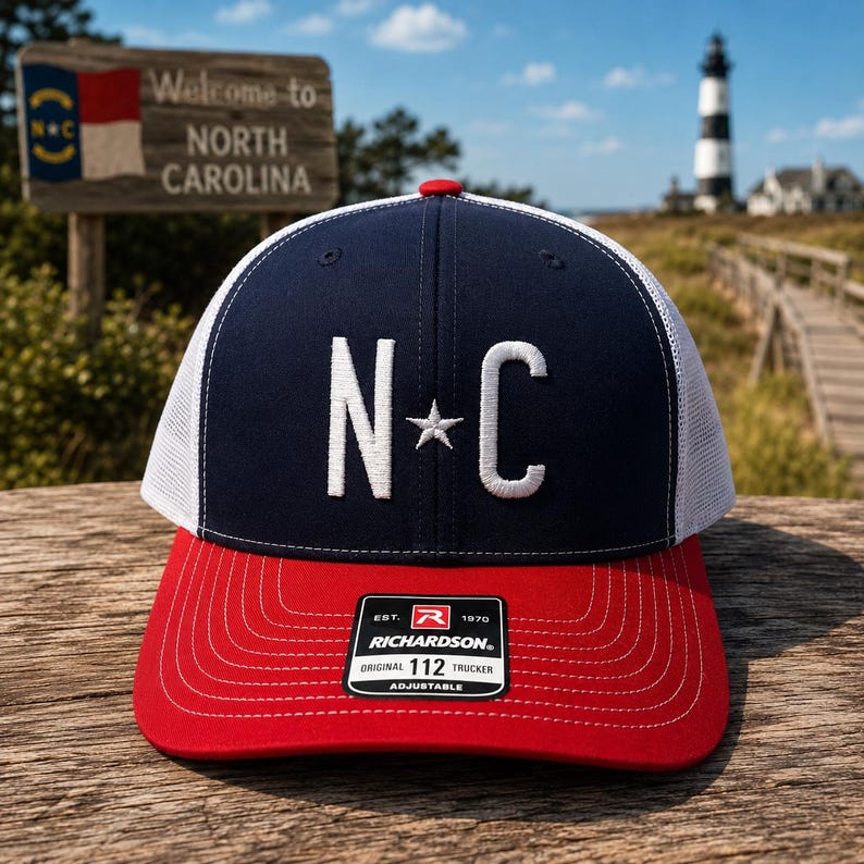 May include: A navy blue and red trucker hat with the letters "NC" and a star embroidered on the front. The hat has a white mesh back and a red brim. The hat has a "Richardson" tag.