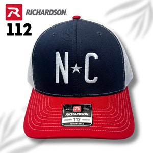 May include: A Richardson 112 trucker cap with a navy blue front and a red peak. The cap has white embroidered "N C" letters and a star. It has a white mesh back and is adjustable. The brand name and logo are visible.