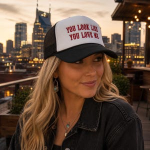 May include: A trucker hat with a white front panel, black mesh back, and a black brim. The front panel features the red embroidered text "YOU LOOK LIKE YOU LOVE ME" in a bold, sans-serif font. The hat has a structured crown and an adjustable strap.