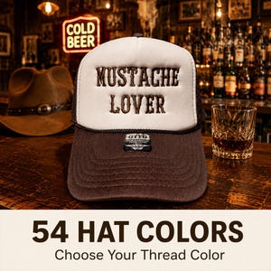 May include: A white and brown trucker hat featuring the embroidered text MUSTACHE LOVER on the front foam panel. The hat has a dark brown brim and a black rope detail across the base of the white front panel, with an Otto brand label attached.