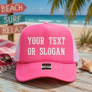 May include: A vibrant pink trucker hat with a white mesh back and a curved brim. The hat features the text "YOUR TEXT OR SLOGAN" in white block letters. A small "OTTO" label is visible on the front.