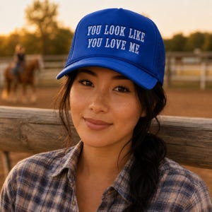 May include: A bright blue trucker hat featuring a mesh back and a solid front panel. The text YOU LOOK LIKE YOU LOVE ME is embroidered in white capital letters across the front of the cap. The hat has a structured crown and a curved brim.