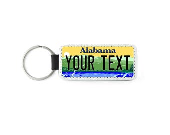 May include: A white metal keychain with a black strap. The keychain features a yellow and blue Alabama license plate design with the text "YOUR TEXT" in black.