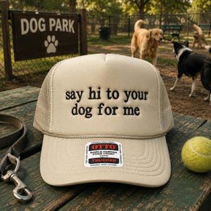 May include: Tan trucker hat with the text "say hi to your dog for me" embroidered in black. The hat has a mesh back and a curved brim. A tennis ball and a dog leash are nearby.