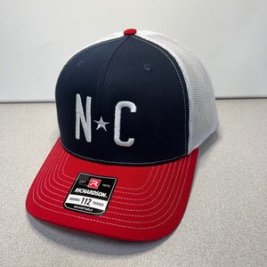 May include: A navy blue and red trucker cap with a white mesh back. The front features the letters "NC" with a star, embroidered in white. The brim is red, and the cap has an adjustable strap.