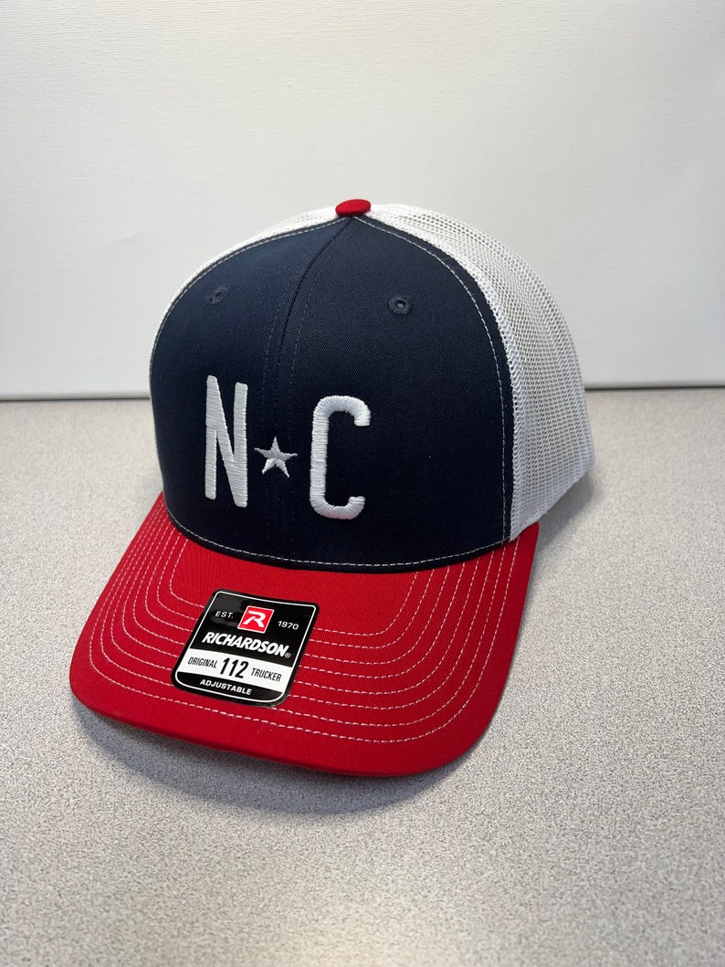 May include: A navy blue and red trucker cap with a white mesh back. The front features the letters "NC" with a star, embroidered in white. The brim is red, and the cap has an adjustable strap.