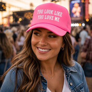 May include: A bright pink trucker hat featuring the white text "YOU LOOK LIKE YOU LOVE ME" printed on the front panel. The cap has a structured foam front, a curved brim, and a mesh back panel, designed with a classic adjustable snapback closure.