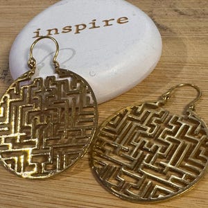 May include: Two gold-colored, openwork earrings with a maze-like design. The earrings are circular and have a hook closure. A white stone with the word "inspire" in gold lettering is in the background.