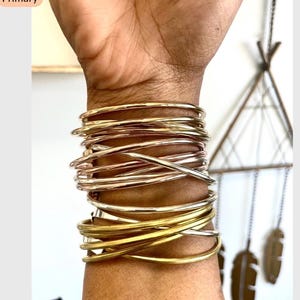 May include: A collection of gold, silver, and rose gold bangles stacked on a wrist. The bangles vary in thickness and finish, creating a layered effect. The bracelets are a mix of shiny and matte textures.