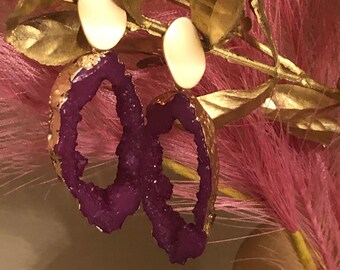 May include: A pair of gold earrings with purple agate stone accents. The earrings are oval shaped and have a gold rim.
