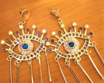 May include: A pair of gold-tone earrings with a blue evil eye design. The earrings are adorned with purple and clear rhinestones and have dangling gold chains.