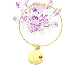 May include: Gold bangle bracelet with a hammered circle charm featuring a blue evil eye. The bracelet is laying on top of a purple crystal lotus flower.