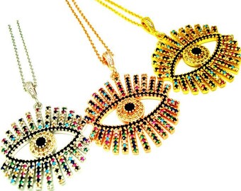 May include: Three eye-shaped pendant necklaces. Each pendant features a central black pupil surrounded by colorful gemstones and a gold or silver-toned frame. The necklaces hang from delicate chains, with a white background.