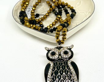 May include: A black and gold beaded necklace with a large black owl pendant adorned with rhinestones. The necklace is displayed on a white heart-shaped dish.