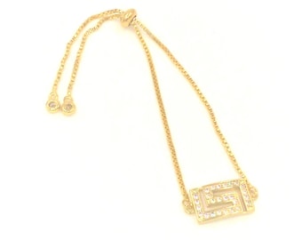 May include: A gold-colored bracelet with a geometric design. The bracelet features a rectangular centerpiece with a maze-like pattern, encrusted with small, clear crystals. The adjustable chain has small, round, crystal-studded accents at the ends.