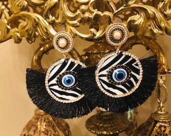 May include: A pair of black and white zebra print earrings with a blue evil eye charm and black fringe. The earrings are gold-toned and have a pearl-like bead at the top.