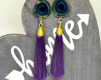 May include: A pair of earrings with a gold-toned metal setting and a green, blue, and black eye-shaped resin pendant. Each earring has a long purple tassel.