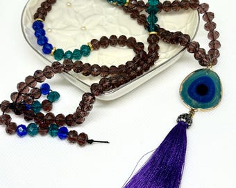May include: A beaded necklace with a purple tassel and an eye-shaped pendant. The necklace features brown, blue, and green beads. The pendant has a blue and green design with a gold-colored border. The tassel is a deep purple color.