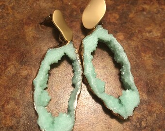 May include: A pair of gold and light green agate earrings. The earrings are oval shaped with a gold backing and a light green agate stone with a crystal-like texture.