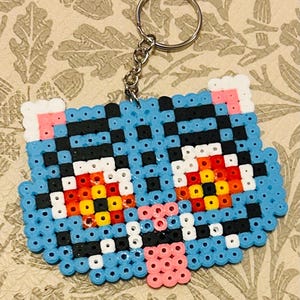 May include: A blue cat-shaped keychain crafted from colorful plastic beads. The cat features black, white, pink, yellow, and red details, with a silver keyring attached. The design is pixelated, giving it a retro aesthetic.