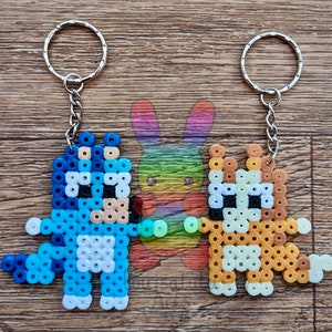 May include: Two beaded keychains, one blue and one orange, shaped like cartoon animals. The blue keychain is shaped like a dog with a white belly and a long tail. The orange keychain is shaped like a cat with a white face and a long tail.