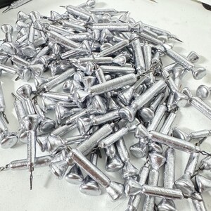 May include: A pile of silver-colored fishing weights. Each weight has a cylindrical body with a flared base and a pointed tip. The weights are scattered across a light-colored surface, with some connected by small metal loops.