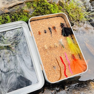 May include: A metal fly fishing box with a cork interior. The box is filled with a variety of colourful flies and fishing lures.