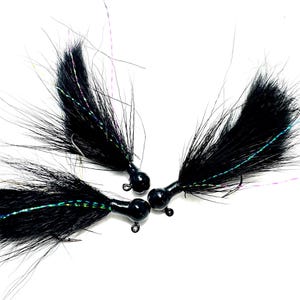 May include: Three black fishing jigs with long, feathery tails and iridescent accents. Each jig has a round, black head and a small hook. The jigs are arranged against a white background, showcasing their design.