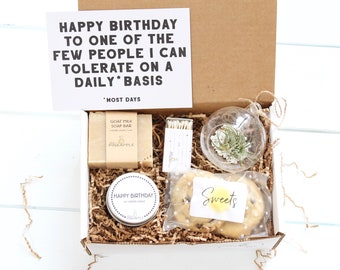 May include: A brown cardboard gift box filled with a variety of items, including a candle, soap, matches, a small plant in a glass globe, and cookies. The box also includes a card with the text "Happy Birthday to one of the few people I can tolerate on a daily basis* *Most days".