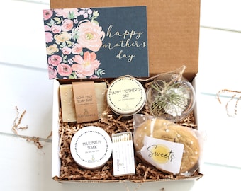 May include: A gift box filled with items for Mother's Day. The box contains a floral card, a goat milk soap bar, a candle, a small potted plant, a bath soak, matches, and a cookie.