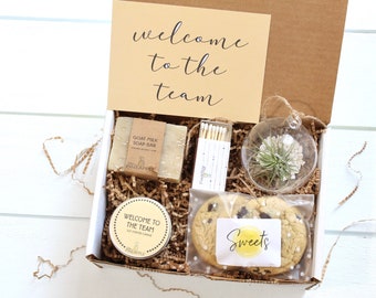 May include: A brown cardboard box filled with a welcome to the team gift set. The box contains a goat milk soap bar, a small jar of scented candle, a glass globe with a small plant, a pack of matches, and a chocolate chip cookie.