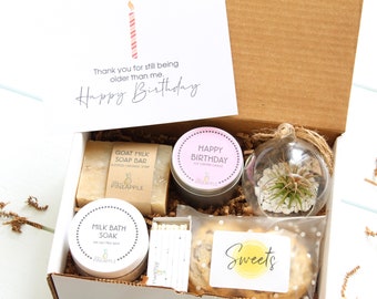 May include: A gift box filled with birthday presents, including a goat milk soap bar, a soy scented candle, a milk bath soak, a small potted plant, and a cookie. The box is decorated with a card that says "Thank you for still being older than me. Happy Birthday."