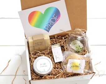 May include: A white gift box filled with a rainbow-themed pride gift set. The box contains a goat milk soap bar, a soy scented candle, a small glass terrarium with a succulent, a pack of matches, and a cookie with a "Sweets" label.