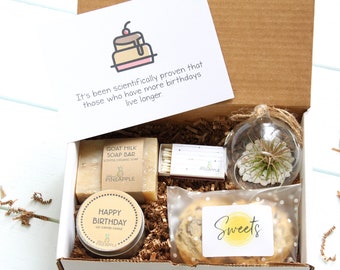 May include: A brown cardboard gift box filled with birthday gifts. The box contains a goat milk soap bar, a candle, a small glass terrarium with a plant, a pack of matches, and a cookie. The card on top of the box reads "It's been scientifically proven that those who have more birthdays live longer."