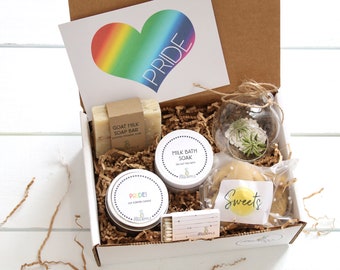 May include: A gift box filled with a variety of items, including a rainbow heart card with the word "PRIDE" on it, a goat milk soap bar, a milk bath soak, a small glass terrarium with a plant, a candle with the word "PRIDE" on it, a box of matches, and a cookie with the word "Sweets" on it.