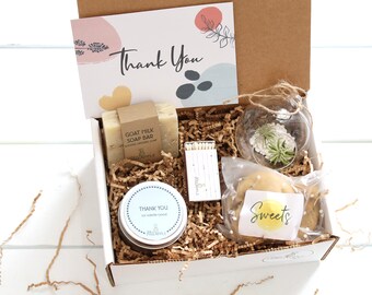 May include: A gift box filled with various items, including a goat milk soap bar, a small potted plant, a candle, matches, and a cookie. The box is lined with brown paper shred and has a card that says "Thank You".