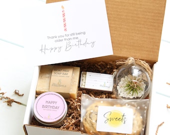 May include: A gift box filled with birthday presents, including a candle, soap, matches, a terrarium, and cookies. The box has a card that says "Thank you for still being older than me. Happy Birthday."